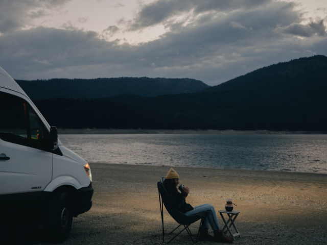 A woman sits in a chair next to a van next to a lake. RV Lifestyle Experts
