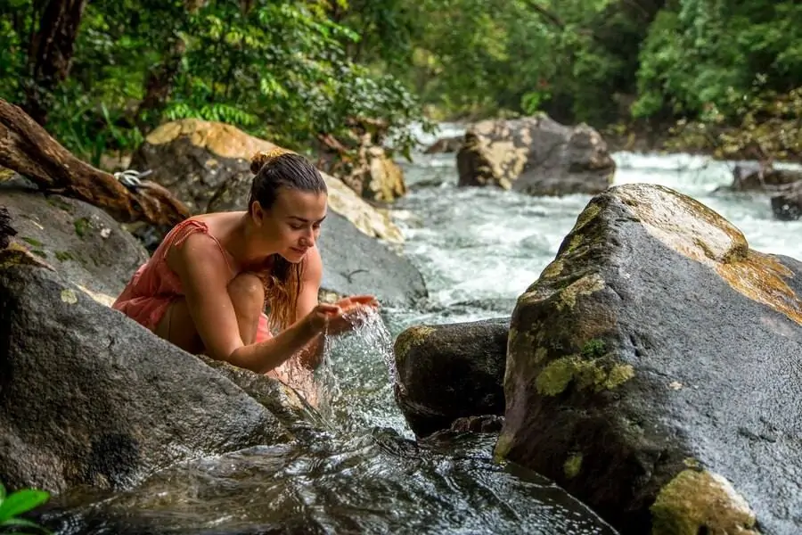 A woman scoops water from a river, surrounded by rocks and lush greenery, with rushing water in the background. RV Lifestyle Experts
