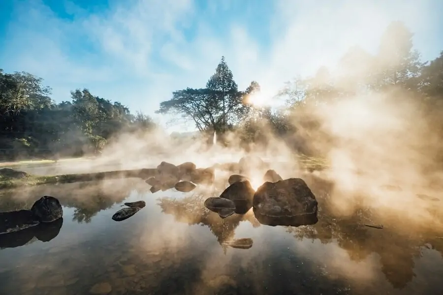 Sunlight peeking through mist above a tranquil hot spring with large stones and reflective clear water surrounded by lush trees. RV Lifestyle Experts
